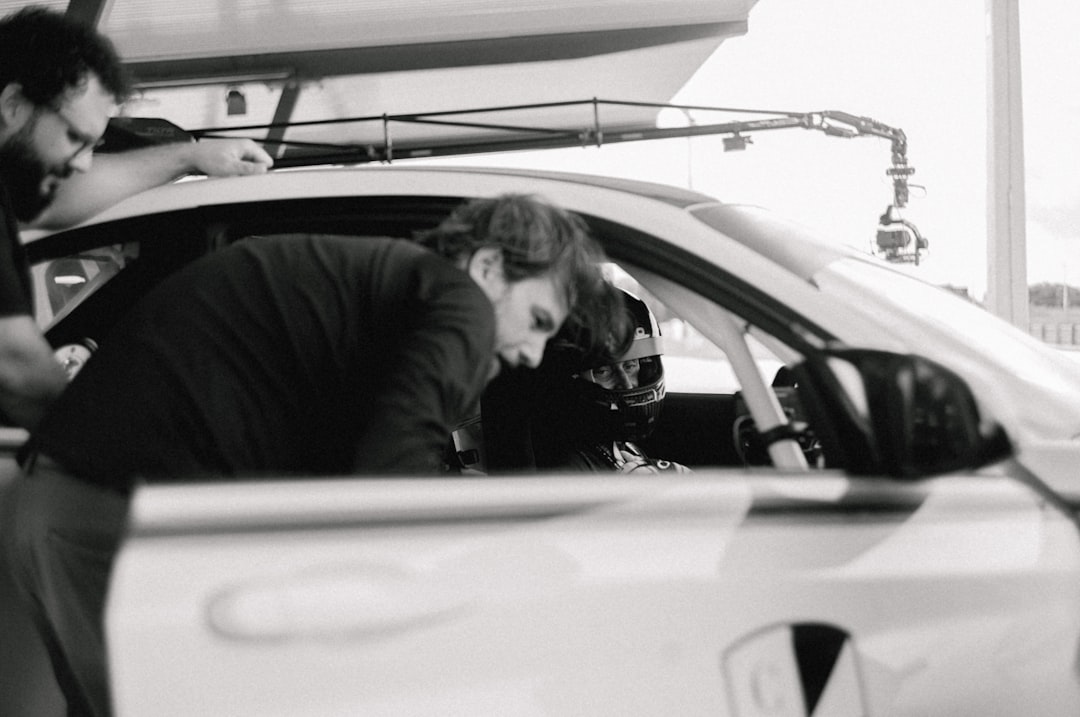 Two men stand beside a car, leaning in to talk to a helmeted driver sitting inside, at what appears to be a racetrack or pit stop.
