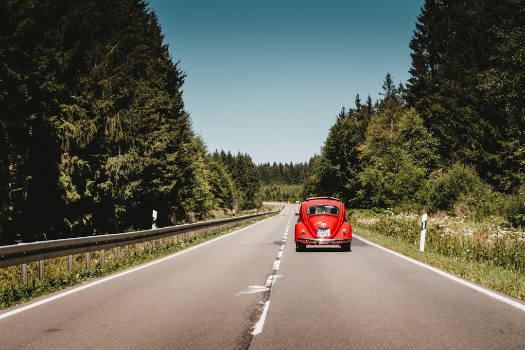 A red vintage car drives down an empty two-lane road surrounded by trees under a clear blue sky. A red vintage car drives down an empty two-lane road surrounded by trees under a clear blue sky.