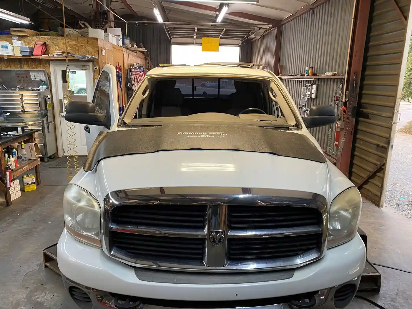 A white Dodge pickup truck with missing windshield and open hood inside an auto repair shop. Tools and equipment are visible on shelves and walls around the workspace.