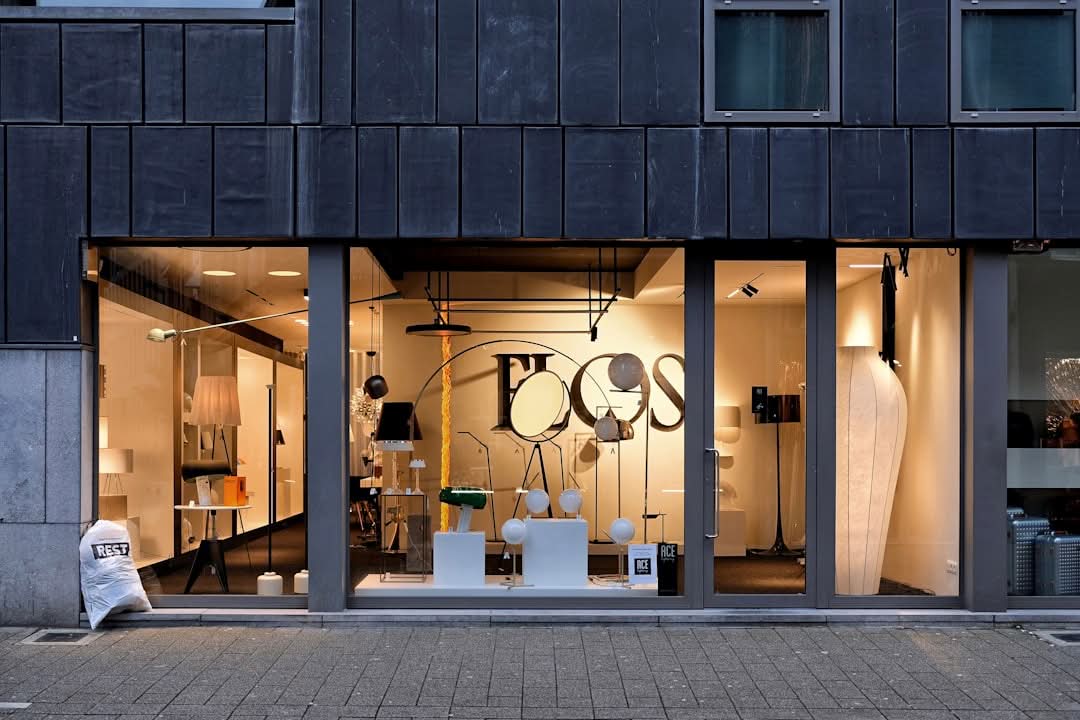 A modern lighting store with large windows displays various lamps and light fixtures inside. A bag sits outside on the sidewalk. A modern lighting store with large windows displays various lamps and light fixtures inside. A bag sits outside on the sidewalk.