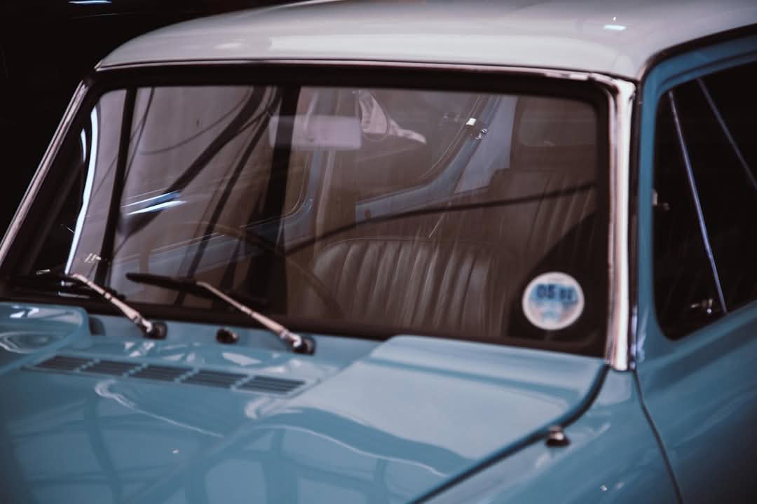 Close-up of the front windshield and interior of a vintage light blue car, showing the steering wheel, dashboard, and a circular sticker on the window.